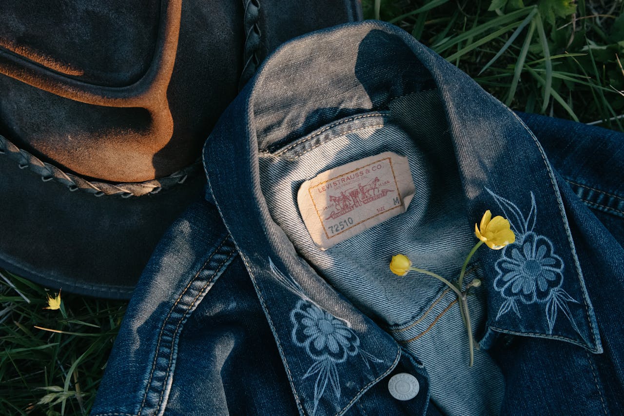 Top view of small yellow fragile flower on denim jacket near hat on green grass in daytime