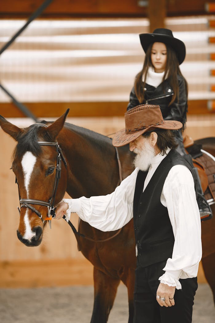 An elderly cowboy assists a young cowgirl riding a brown horse indoors, showcasing equestrian skills.